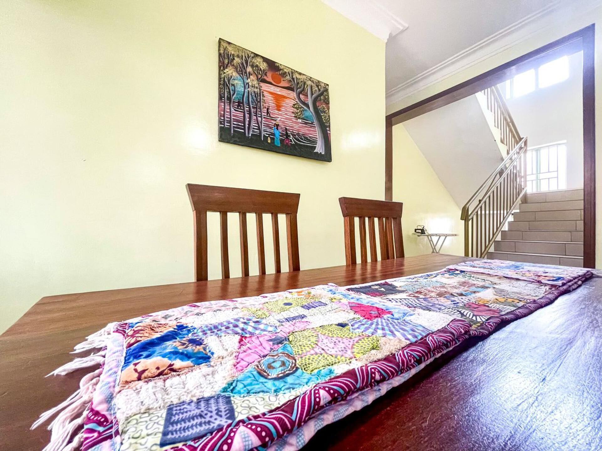 Una mesa de comedor con un colcha colorida, dos sillas de madera y un cuadro en la pared.