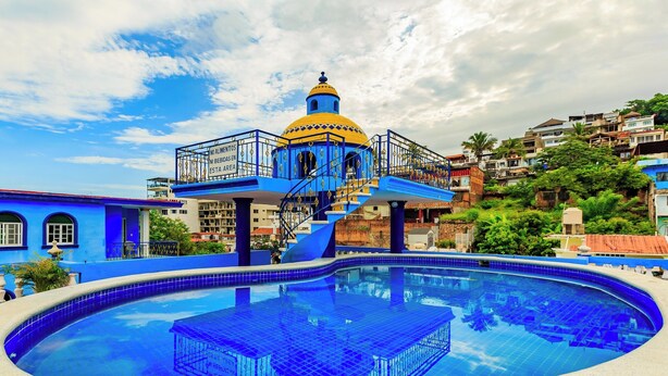 A large swimming pool with a blue tiled deck and a central structure with a yellow dome. - OYO Casa Mission (Puerto Vallarta)
