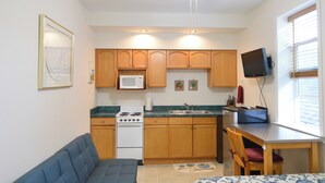 A compact kitchen with wooden cabinets, a microwave, a stove, and a dining area with a table and chairs. - Historic Apartment in Downtown Seward (Seward)