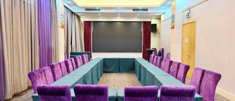 A conference room with a long table and purple chairs, a projector screen, and a stage area.