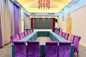 Un salón de conferencias con una larga mesa y sillas moradas, una pantalla de proyector y un escenario.