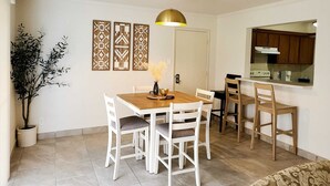 Uma sala de jantar com mesa de madeira e cadeiras brancas, um vaso com plantas secas e uma luminária pendente.