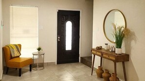 Um corredor com porta, uma pequena mesa com uma planta, uma cadeira e um aparador com espelho e vasos.