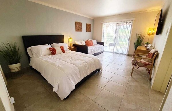 A hotel room with a large bed, a smaller bed, a desk with a chair, a TV mounted on the wall, and a sliding glass door leading to a balcony.