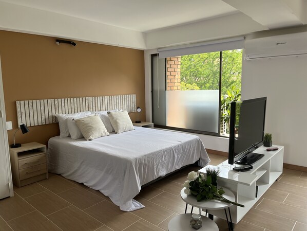 Un dormitorio moderno con una cama grande, un televisor, una mesita con flores y un ventanal con vistas al verdor.