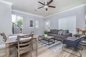 Un salón espacioso con un sofá gris, una mesa de centro y un sillón azul. Hay una zona de comedor con una mesa y sillas de madera.