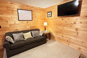 Een kamer met houten paneelwanden, een donkere bank met kussens, een kleine tafel met een lamp en een flatscreen televisie die aan de muur is bevestigd.