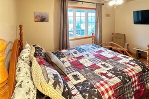 Ein Schlafzimmer mit einem hölzernen Bett, einer Steppdecke, einem Fenster mit Vorhängen, einem Sessel und einem an der Wand befestigten Fernseher.