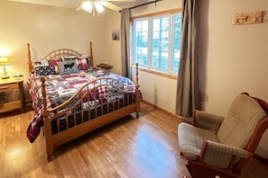 Une chambre à coucher avec un lit en bois, une chaise, une table de chevet, une fenêtre avec des rideaux et un tableau accroché au mur.