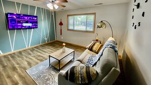 Un salon doté d’un ventilateur de plafond, d’un téléviseur à écran plat fixé au mur, d’une fenêtre avec des stores, d’un canapé agrémenté de coussins à motifs et d’une petite table sur laquelle est posé un vase.