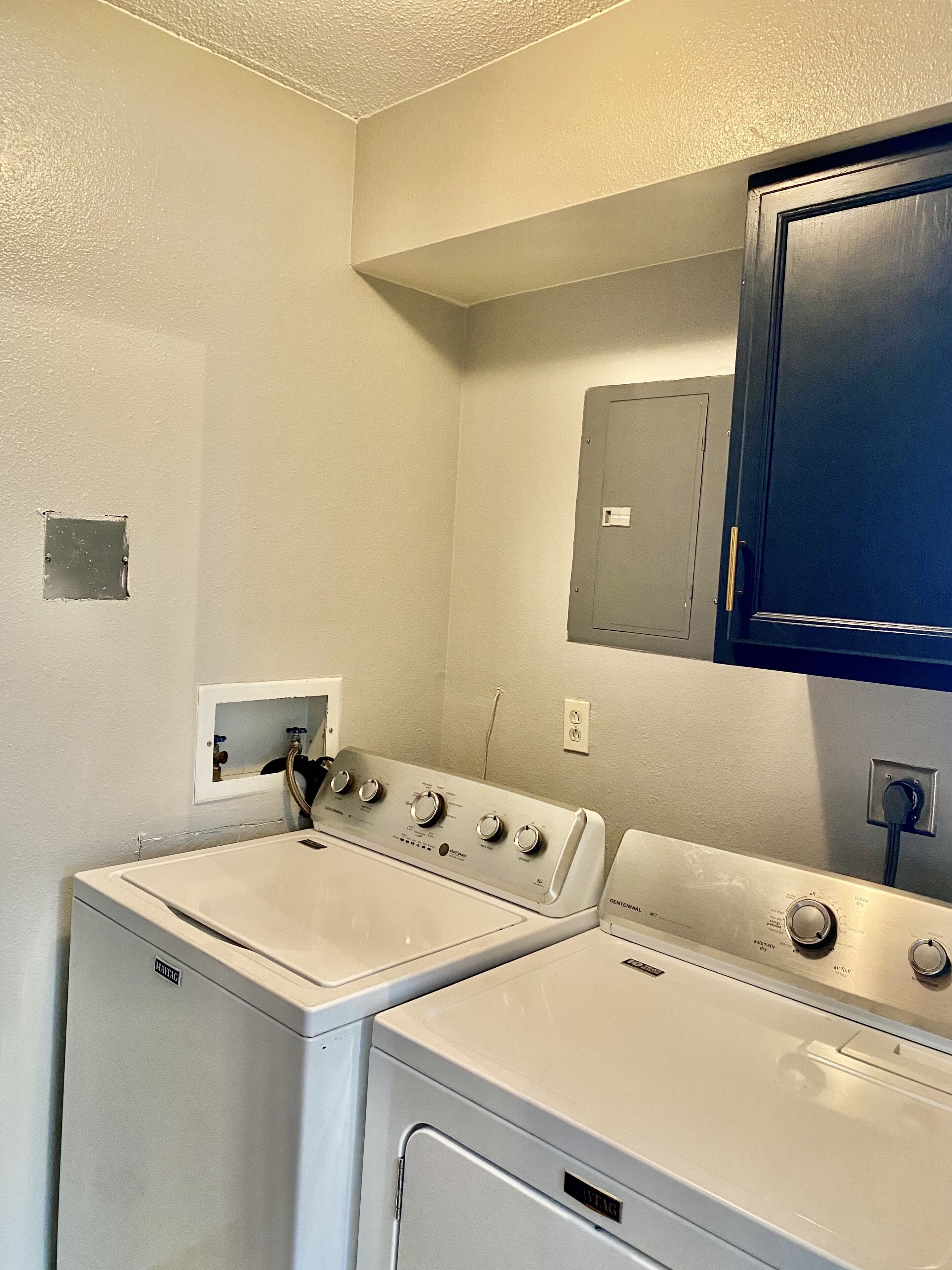 A laundry room with two white front-loading washers and dryers, a small electrical panel, and a blue cabinet above the dryers.