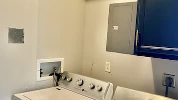 A laundry room with two white front-loading washers and dryers, a small electrical panel, and a blue cabinet above the dryers.