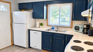 A modern kitchen with a white refrigerator, dishwasher, and stove, blue cabinets, and a window with blinds. - Remodeled apartment in central location (Council Bluffs)
