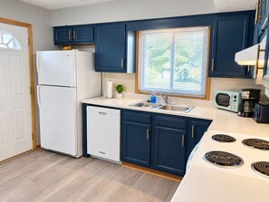 A modern kitchen with a white refrigerator, dishwasher, and stove, blue cabinets, and a window with blinds. - Remodeled apartment in central location (Council Bluffs)