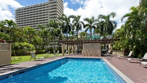 Une piscine entourée de chaises longues et agrémentée d’une pergola, avec des palmiers tout autour et un grand immeuble en arrière-plan.