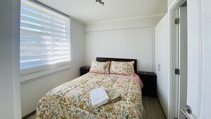 A bedroom with a floral bedspread, a wooden headboard, a nightstand, and a window with blinds.