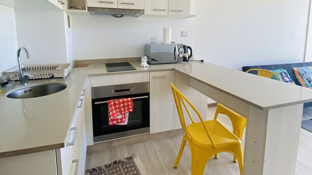 A compact kitchen with white cabinets, a built-in oven, a microwave, and a sink. There is a yellow chair and a patterned rug on the floor.