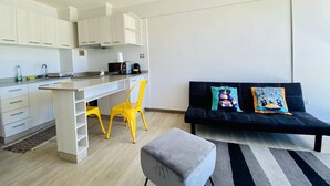 A modern kitchen with white cabinets, a black countertop, and a microwave. There is a yellow chair and a black sofa with colorful cushions.