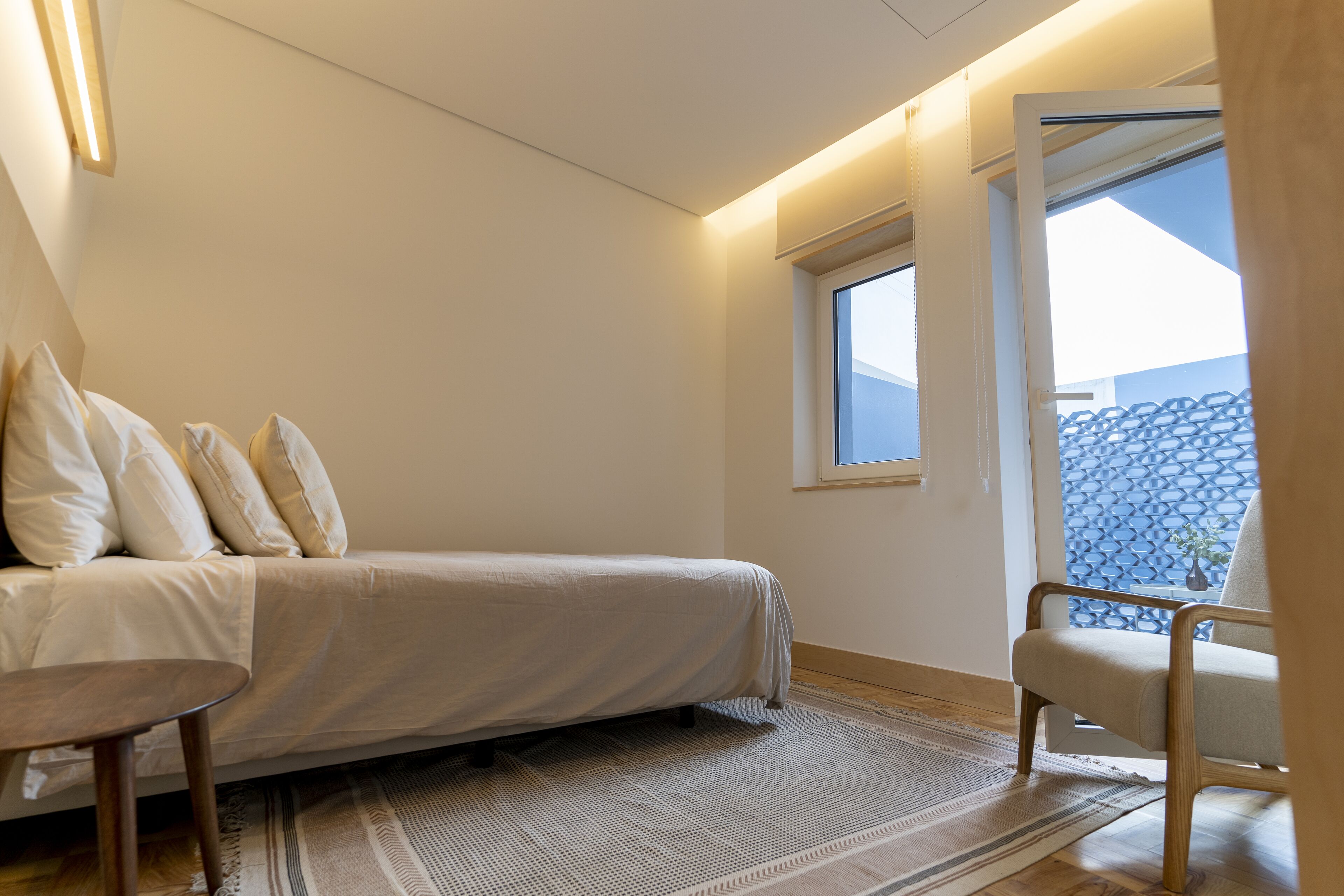 Une chambre à coucher comprenant un lit, une chaise, une petite table et une fenêtre donnant sur un mur bleu à motifs.