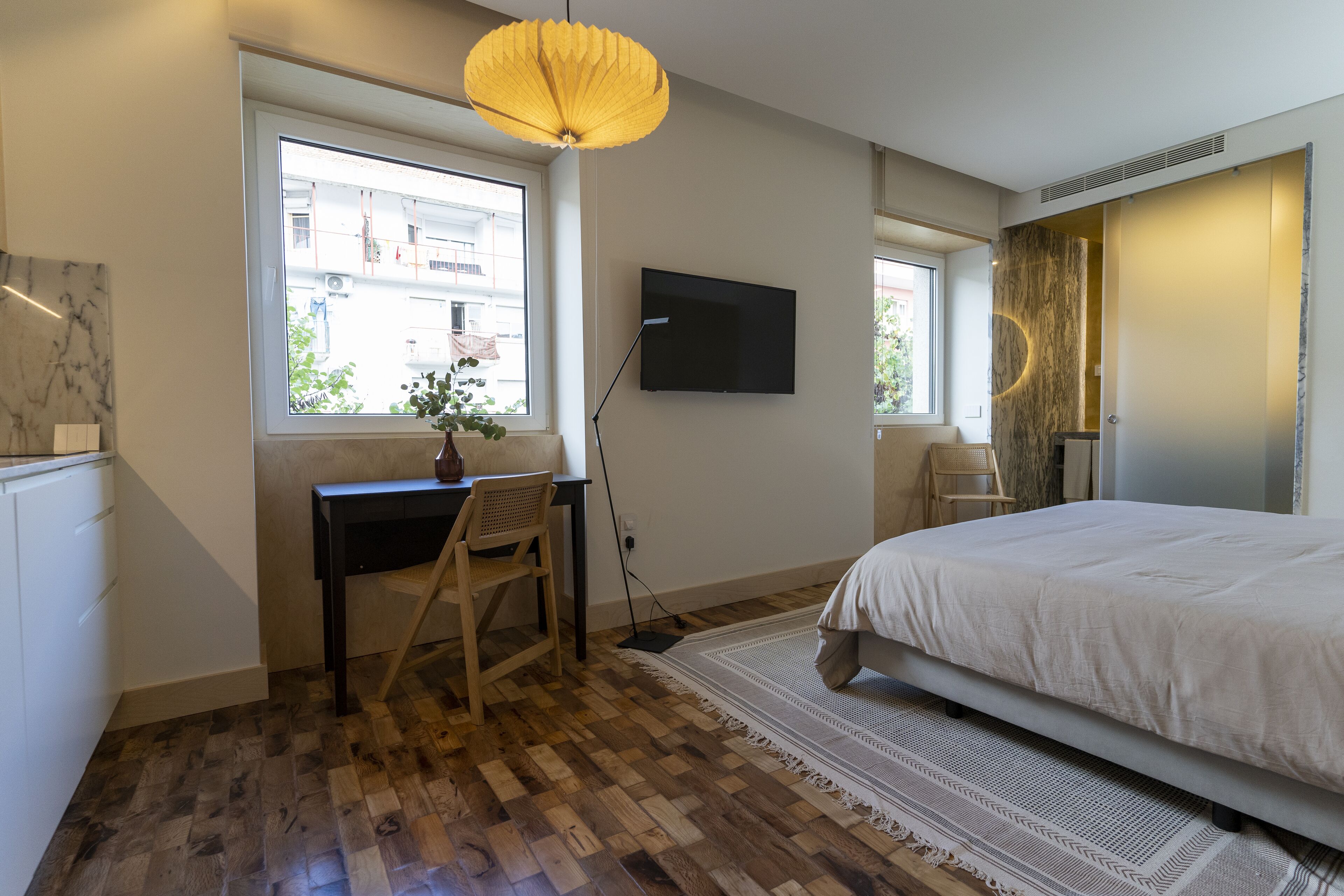 A bedroom with a bed, a desk, a chair, a TV, and a window with a view of buildings.