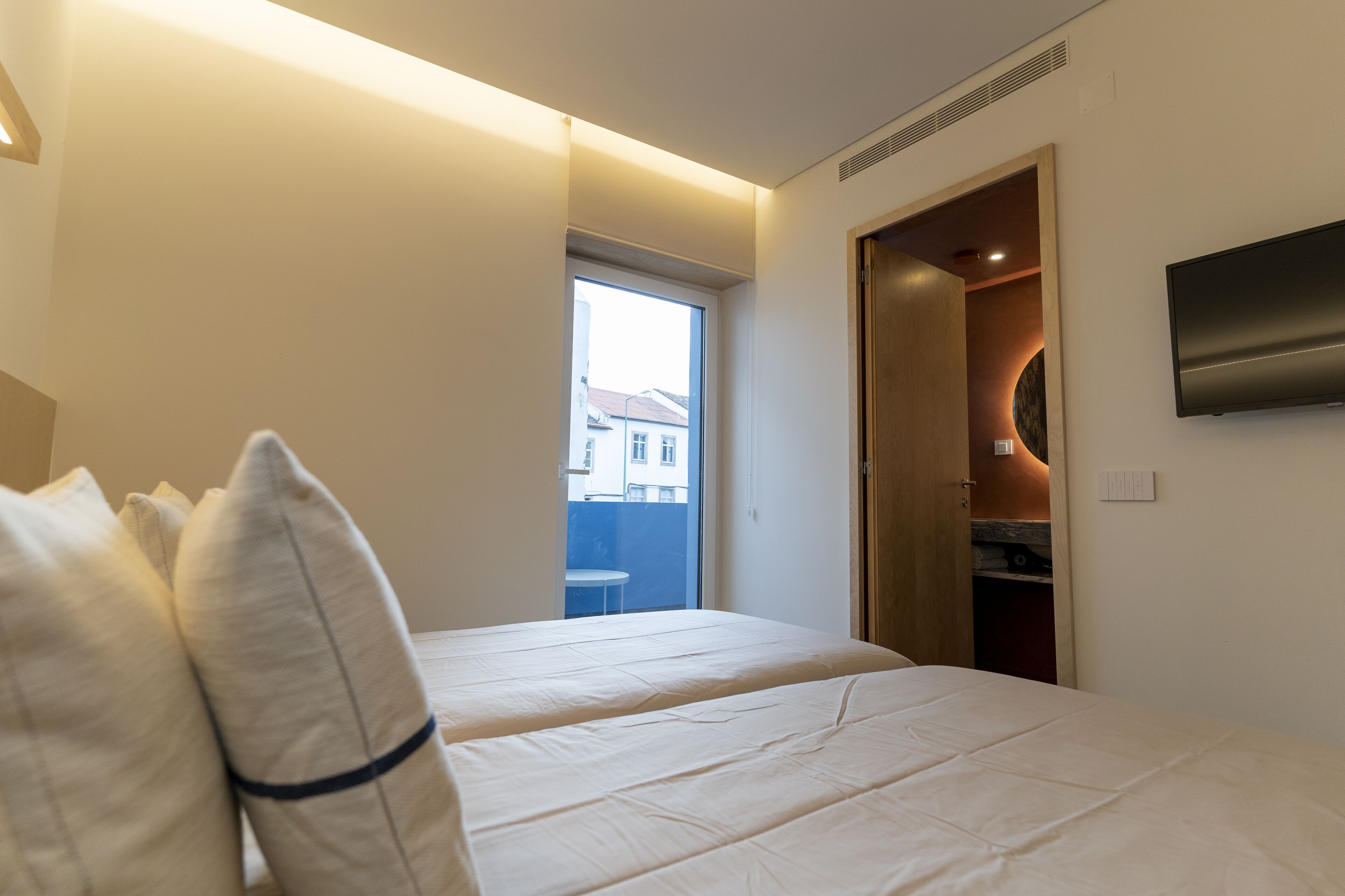 A hotel room with two beds, a TV mounted on the wall, and a view of the outside through a window.