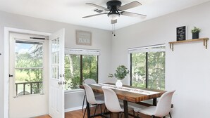 Comedor con una mesa de madera, cuatro sillas, un ventilador de techo y ventanas amplias con vistas a la vegetación exterior.