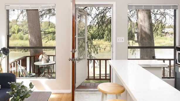 Una cocina moderna con encimera blanca y taburetes de madera, ventanas amplias con vistas a un lago y árboles, y una puerta que da a una terraza exterior.