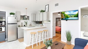 Una cocina moderna con armarios blancos, una isla central con taburetes, un ventilador de techo y un televisor que muestra un paisaje pintoresco.