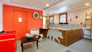 Zona de recepción con mostrador de madera, un reloj, un tablón de anuncios, pared roja y una pequeña mesa con una cesta.