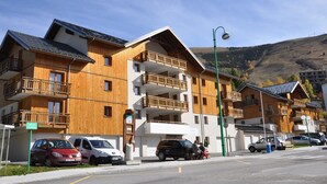 Una hilera de edificios de madera de varios pisos con balcones, coches aparcados y un cielo despejado.