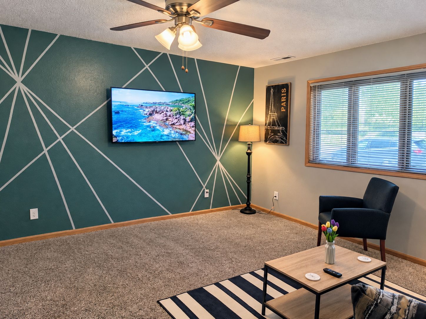 A living room with a television, a ceiling fan, a lamp, a chair, a coffee table, and a window with blinds.