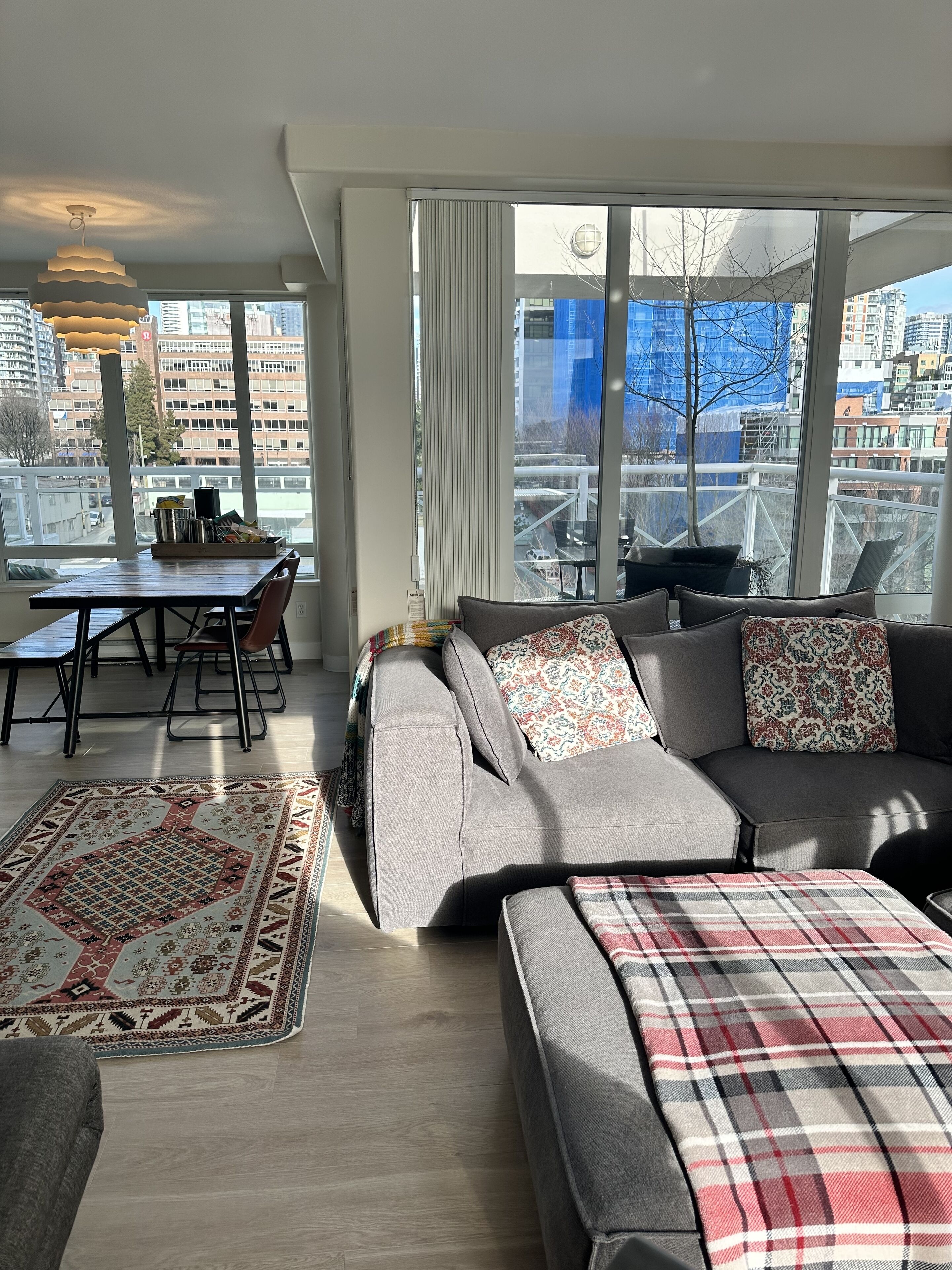 Een moderne woonkamer met een grijze bank, een geruit plaid en een tapijt met een patroon. Er is een zithoek met een tafel en stoelen, en grote ramen met uitzicht op de stad.
