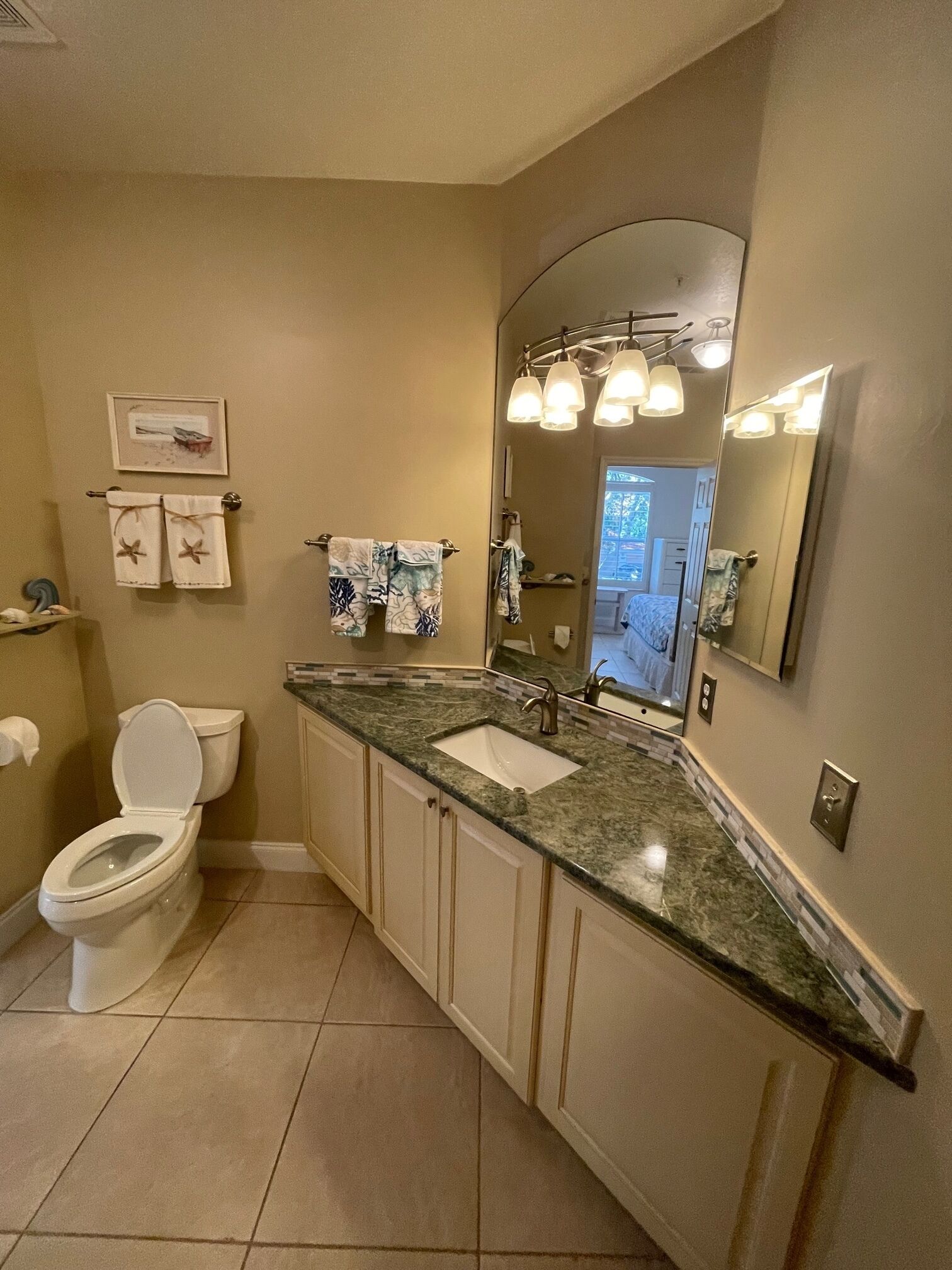 A bathroom with a toilet, sink, and mirror. There are towels hanging on the wall and a picture frame above the toilet.