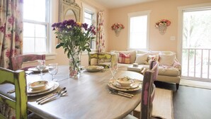Une table à manger dressée avec des assiettes, des verres et des couverts, entourée de rideaux à motifs floraux et d’une chaise ancienne.
