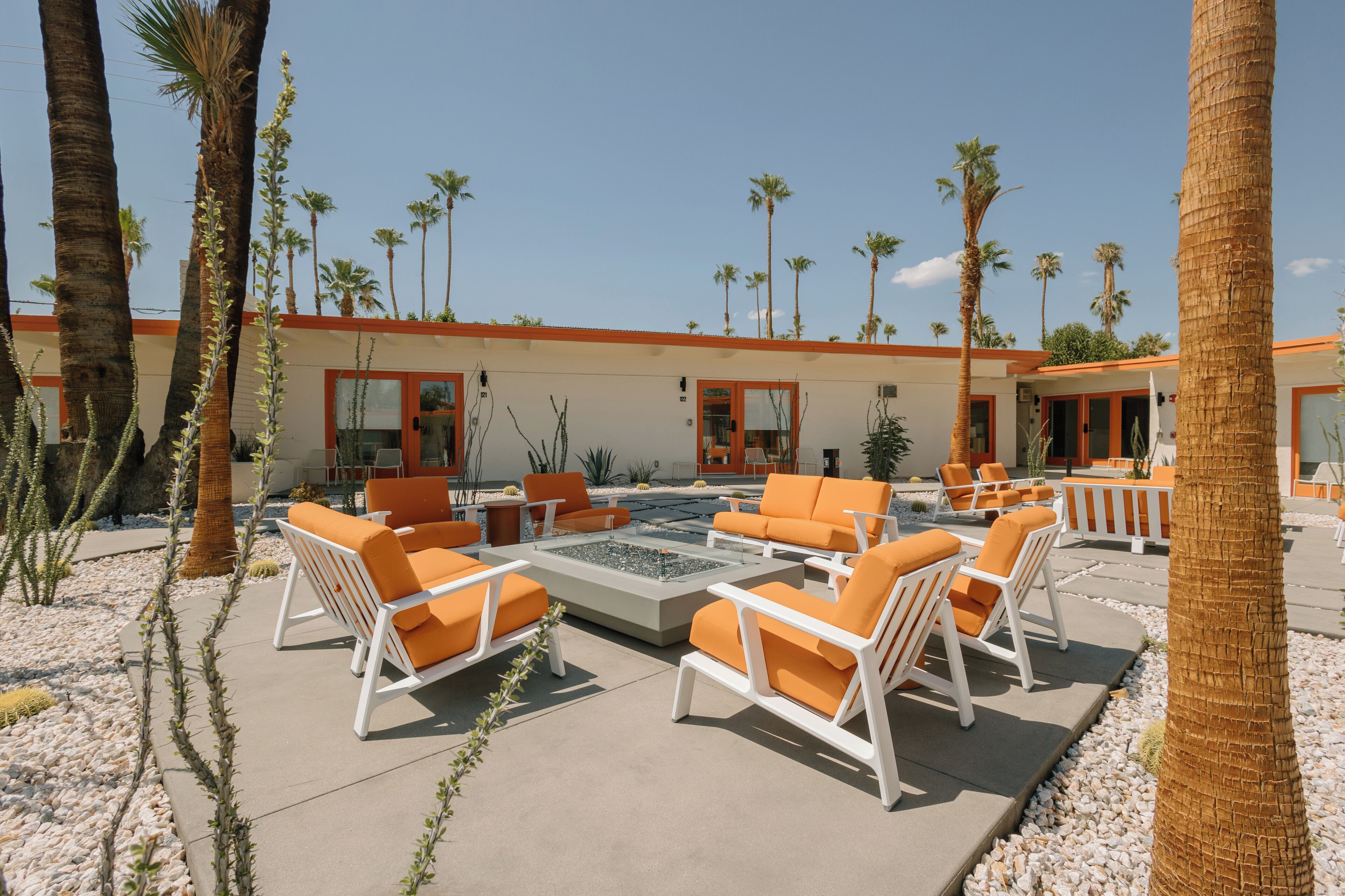 An outdoor lounge area with white and orange lounge chairs, a central fire pit, and palm trees.