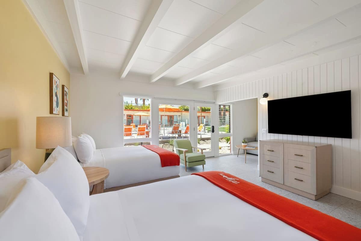 A hotel room with two beds, a TV, a desk, and a view of the outdoor pool area.