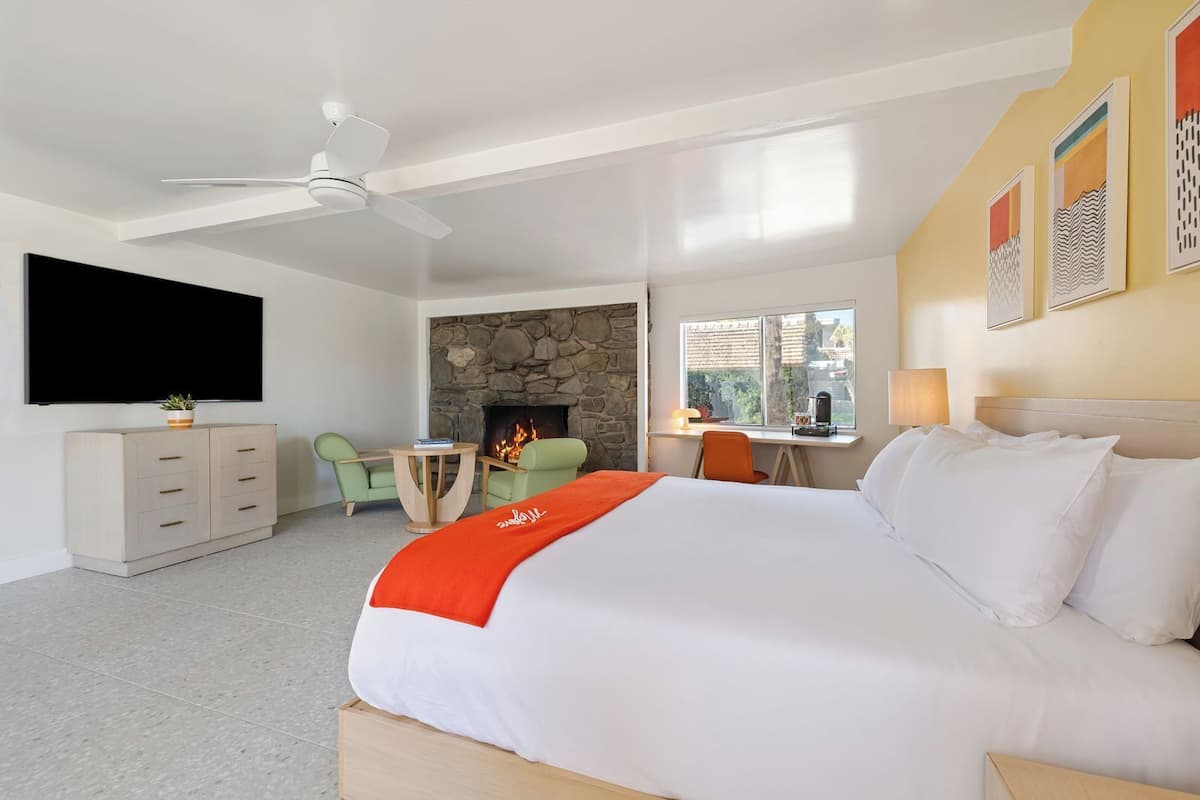 A modern hotel room with a large bed, a flat-screen TV, a stone fireplace, a desk with a chair, and a window with a view.