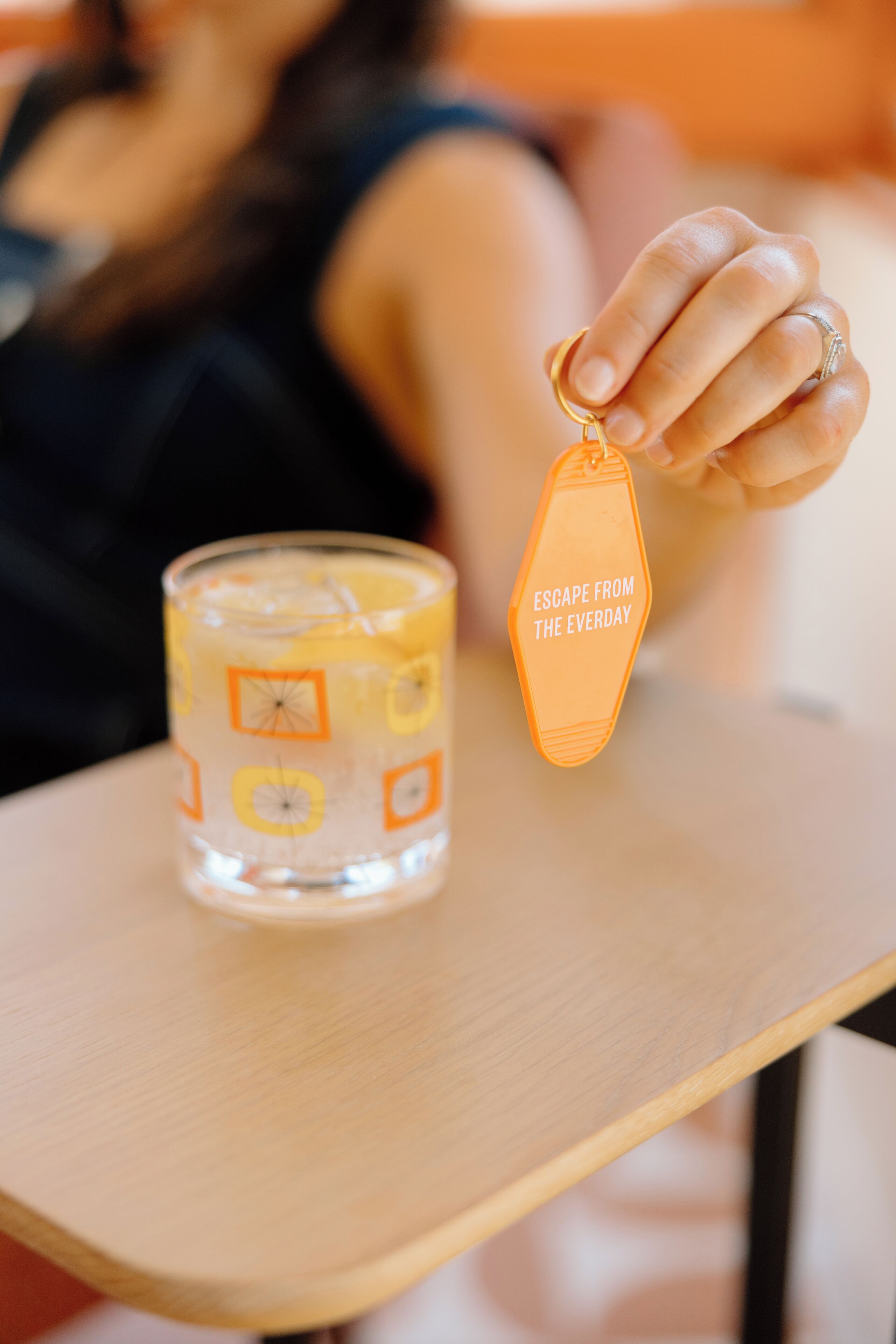 Eine Person hält einen Schlüsselanhänger mit dem Schriftzug "ESCAPE FROM THE EVERYDAY" neben einem Glas mit Zitronenmuster.