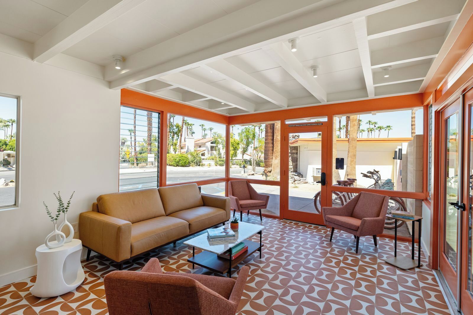 A modern living room with a sofa, armchairs, and a coffee table. Large windows provide a view of the street and palm trees outside.