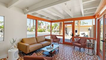 A modern living room with a sofa, armchairs, and a coffee table. Large windows provide a view of the street and palm trees outside.