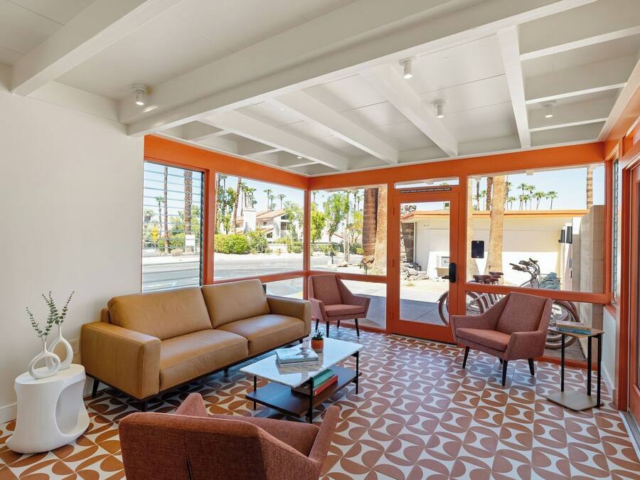 A modern living room with a sofa, armchairs, and a coffee table. Large windows provide a view of the street and palm trees outside.