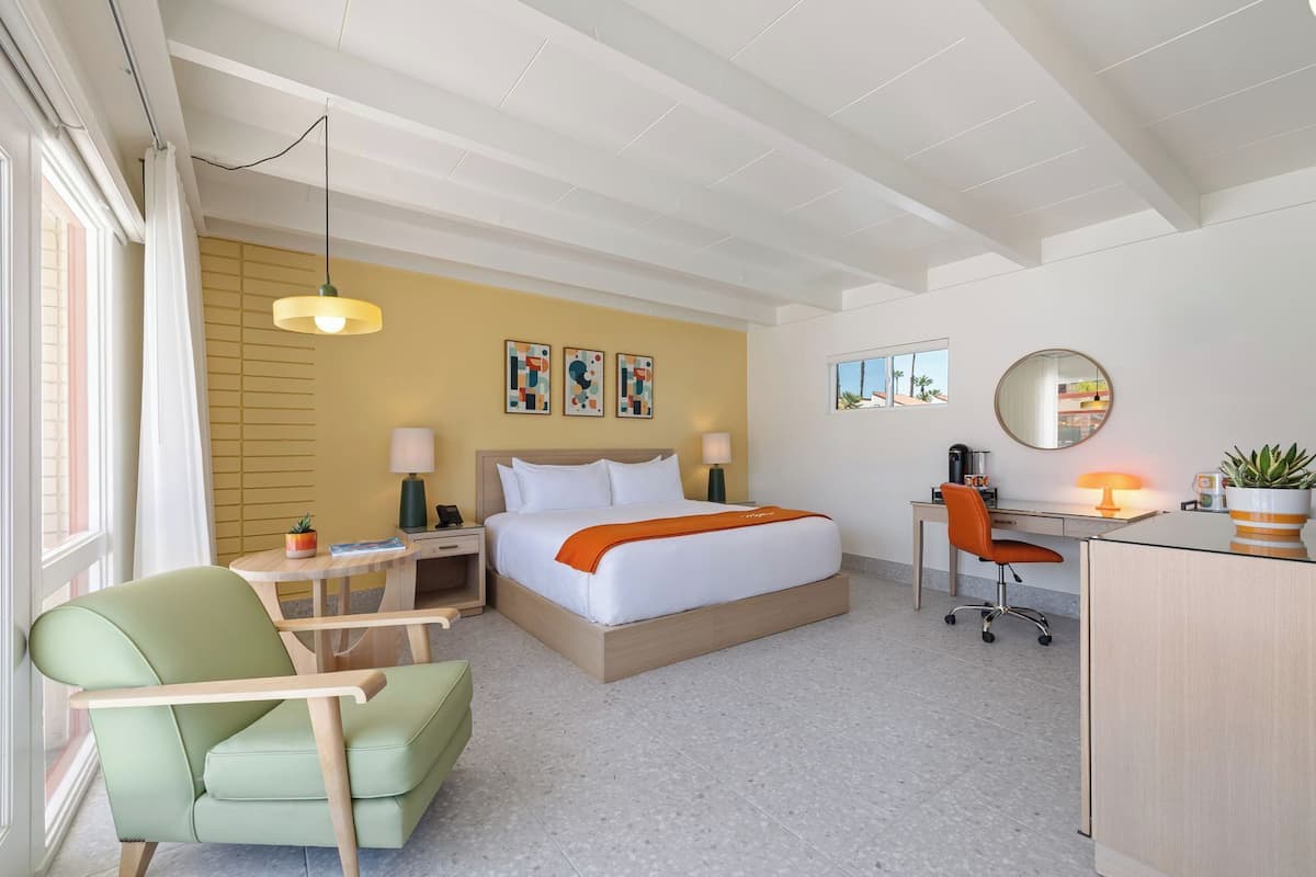 A hotel room with a bed, desk, chair, and a desk lamp.