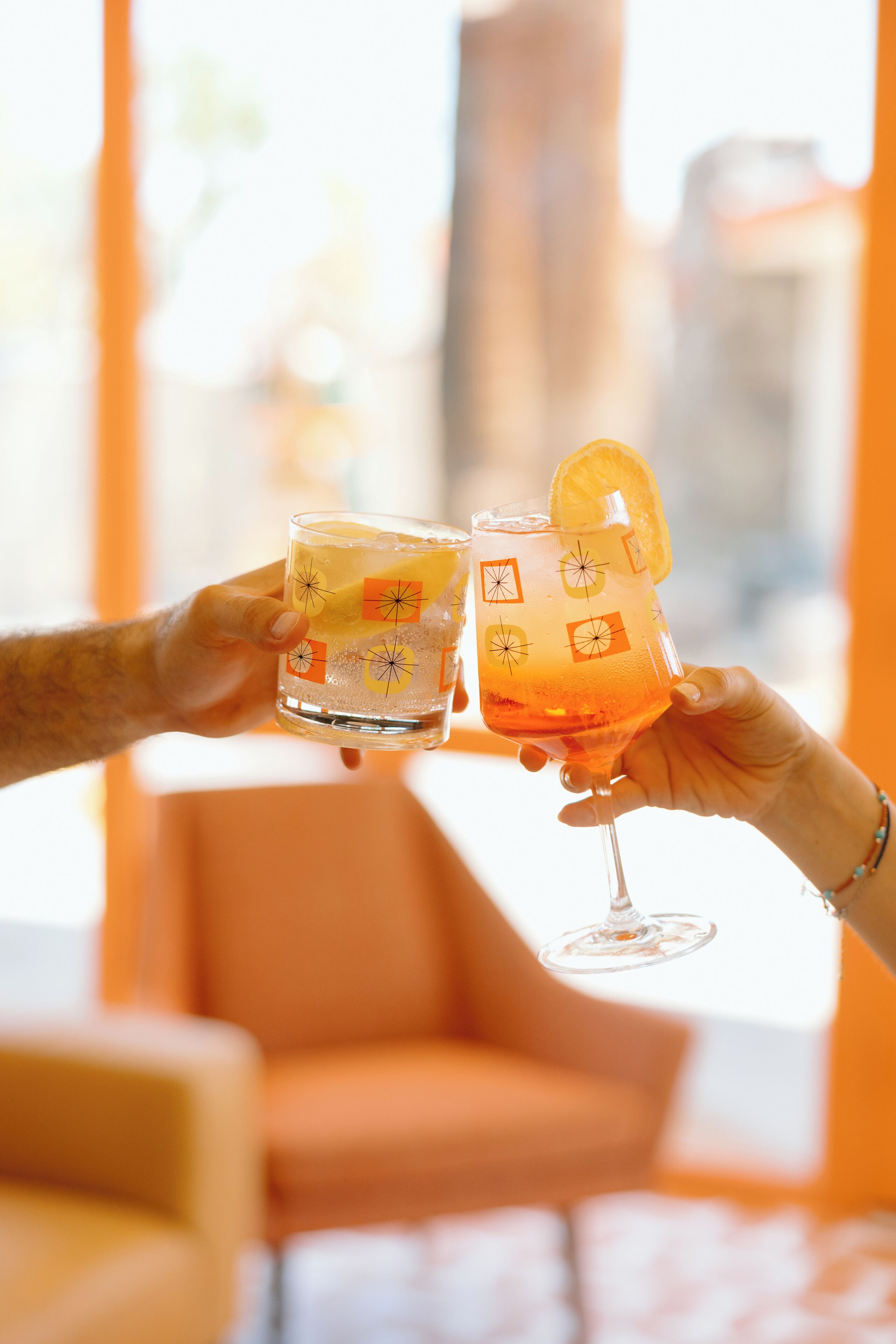 Two people holding glasses with citrus slices, in a room with orange walls and a window.