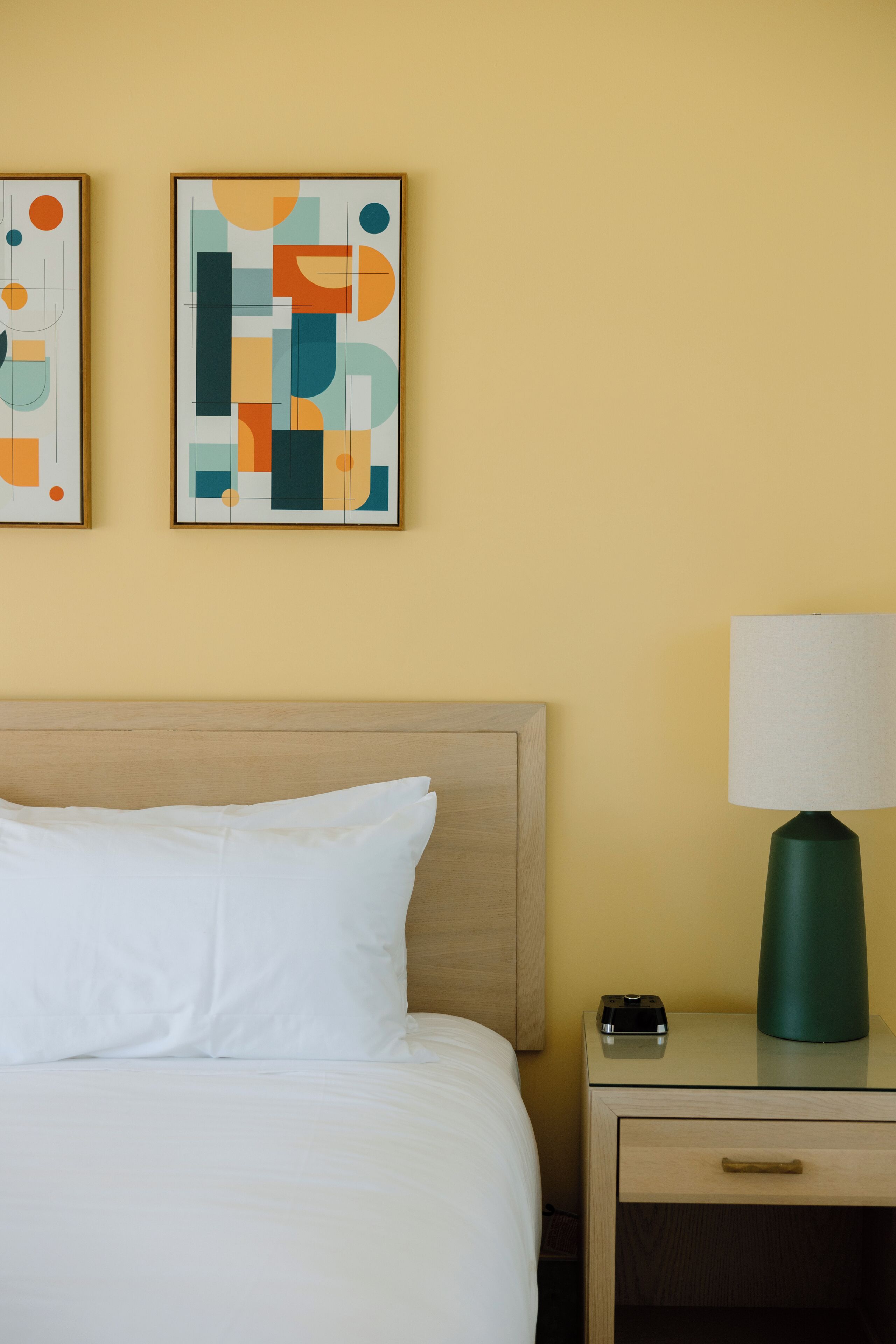 Un lit bien fait, avec du linge de lit blanc, une tête de lit en bois, une table de chevet surmontée d’une lampe verte, et une œuvre d’art abstraite accrochée au mur.