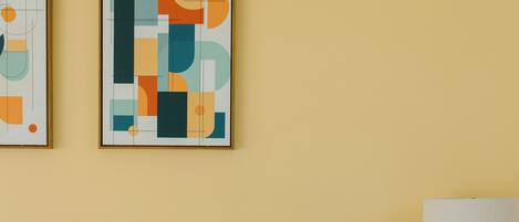 Un lit bien fait, avec du linge de lit blanc, une tête de lit en bois, une table de chevet surmontée d’une lampe verte, et une œuvre d’art abstraite accrochée au mur.
