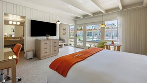 A hotel room with a bed, a TV, a desk, a chair, a dresser, and a view of the pool area. - Mojave Resort - Adults Only (Palm Desert)