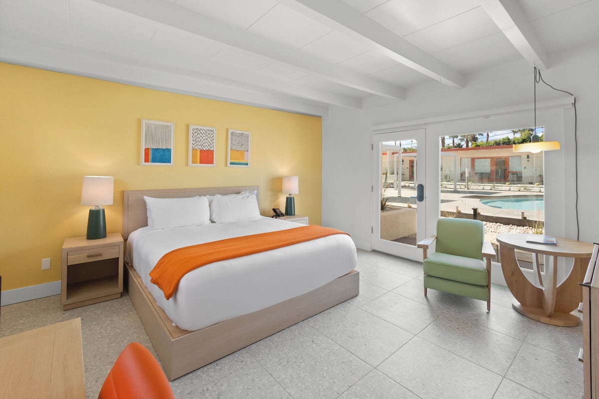 A modern hotel room with a large bed, a green chair, a small table, and a view of a pool through the glass doors.