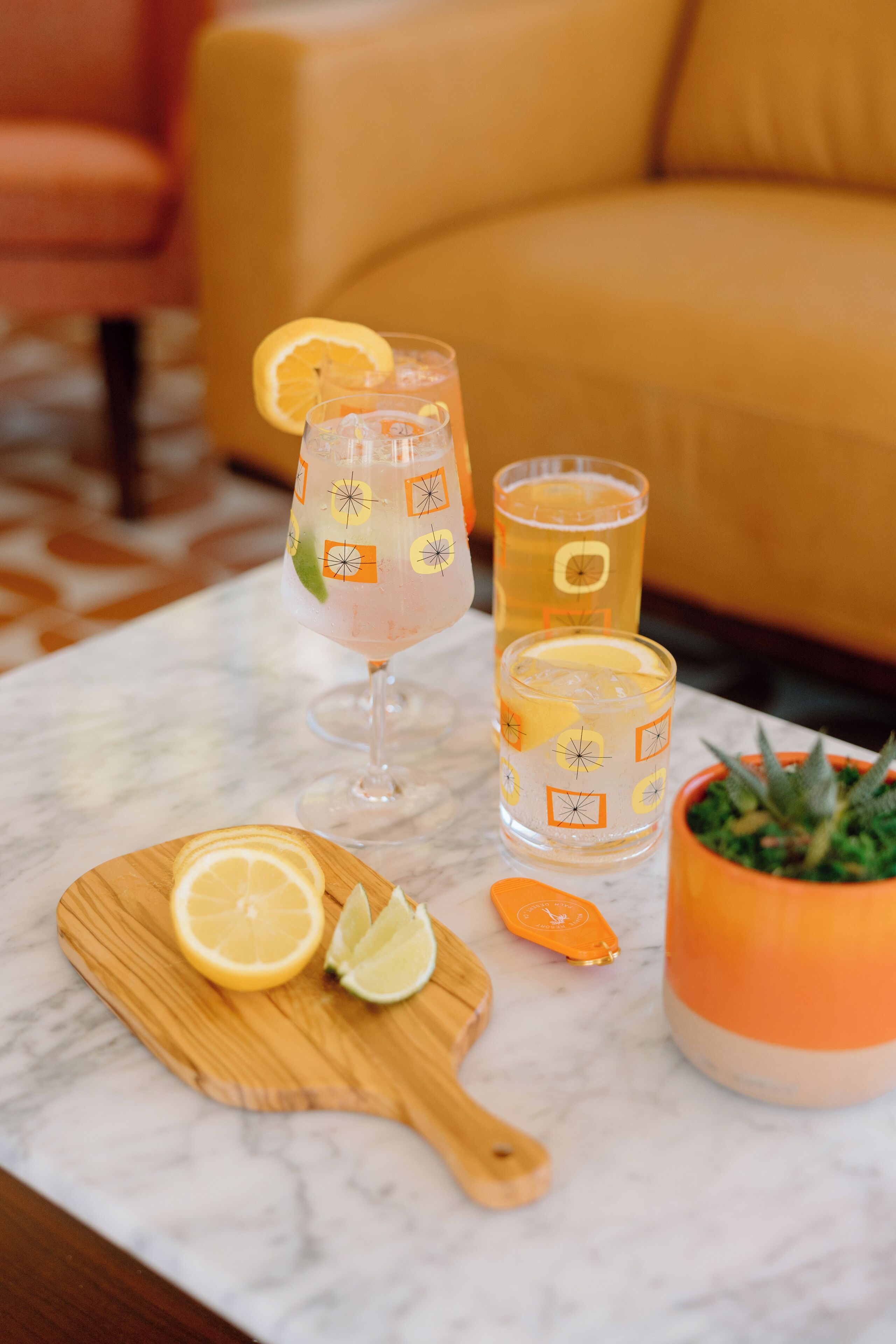 A table with two glasses of a pink beverage, a wooden board with lemon slices, and a small orange pot with a succulent plant.