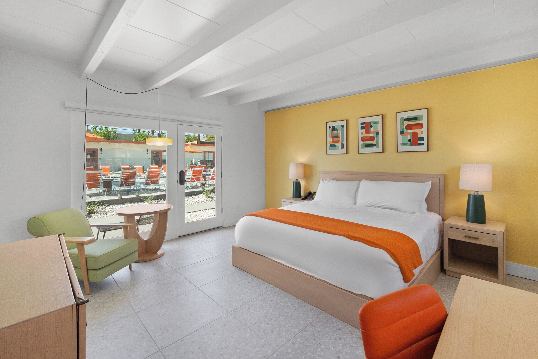 A hotel room with a bed, a desk, a chair, a table, a lamp, and a view of the outside patio.