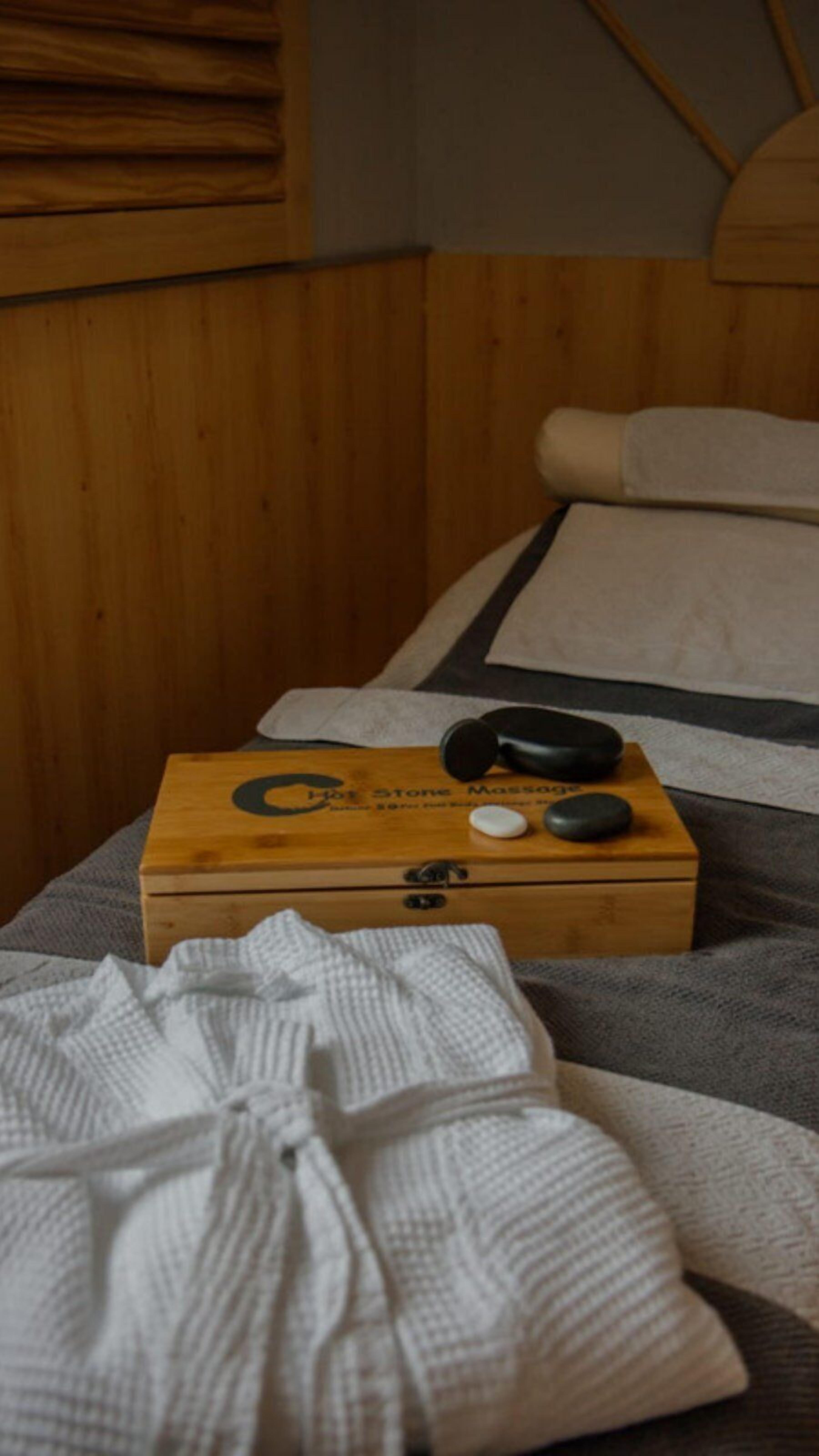 Cabecera de madera, una caja de madera con la etiqueta 'Masaje con Piedras Calientes' y un albornoz blanco sobre una cama con manta a rayas.
